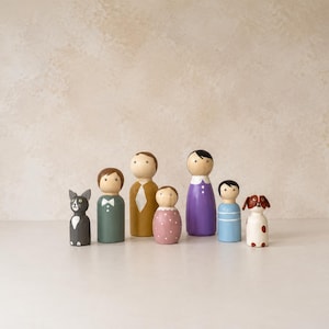 Op de afbeelding: Een set van zeven handbeschilderde houten peg dolls die een familie voorstellen. De figuren bestaan uit een grijze kat, een persoon in een groen pak, een persoon in een beige outfit, een klein persoon in een roze jurk met stippen, een persoon in een paarse jurk, een persoon in een blauw shirt en een bruin-witte hond.