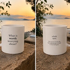Könnte beinhalten: Ein weißer Keramikbecher mit schwarzem Textaufdruck auf beiden Seiten. Eine Seite trägt die Aufschrift "What is worth worrying about?". Die andere Seite zeigt das Wort "scholē" in lateinischen und griechischen Buchstaben sowie die Definition.