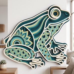 May include: A multi-layered wooden wall art piece shaped like a frog, featuring intricate cutouts and patterns in shades of teal, dark blue, and pale yellow. The design uses stacked layers to create depth, showcasing a mandala-like motif on the body.