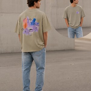 Könnte beinhalten: Ein olivgrünes Kurzarm-T-Shirt mit einer bunten, abstrakten Grafik auf der Rückseite, die ein Gesicht, einen Fisch und japanischen Text zeigt. Auf der Vorderseite befindet sich ein kleines, passendes Motiv auf der Brust. Das T-Shirt wird mit einer hellblauen Jeans kombiniert.