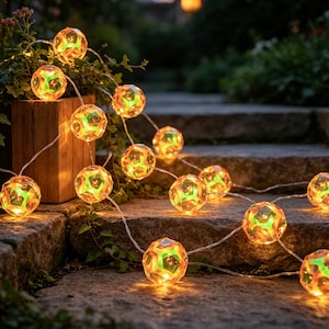 Puede incluir: Una guirnalda de luces LED decorativas con esferas geométricas translúcidas y multifacetadas. Cada orbe emite una luz cálida con tonos verdes y naranjas, conectadas por un cable fino y transparente extendido sobre escalones de piedra.