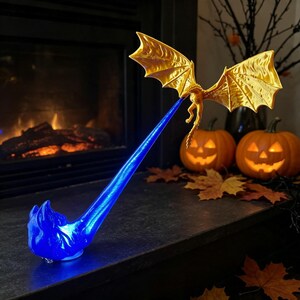 May include: A gold-colored dragon figurine with outstretched wings positioned in the air, emitting a long, translucent blue stream of light or energy that extends downward to a base, creating a dynamic visual effect of a fire-breathing creature.