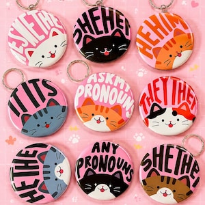 Puede incluir: Nueve llaveros circulares con caras de gatos de dibujos animados y texto de pronombres. Los diseños incluyen gatos con varios colores y patrones de pelaje, cada uno emparejado con letras en negrita como SHE/HER, HE/HIM, THEY/THEM, ASK MY PRONOUNS.