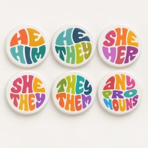 May include: Six circular pin-back badges featuring colourful, wavy typography. The badges display the text: HE HIM, HE THEY, SHE HER, SHE THEY, THEY THEM, and ANY PRONOUNS. Each badge uses a distinct palette of vibrant, multi-coloured letters.