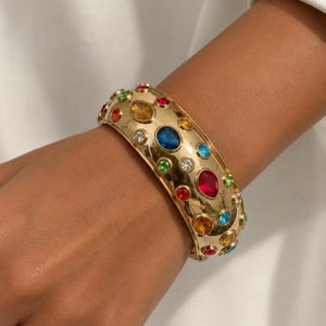 Può includere: Un ampio bracciale rigido in metallo color oro caratterizzato da una serie di pietre preziose sfaccettate multicolori di varie forme e dimensioni, tra cui pietre rosse, blu, verdi, gialle e trasparenti incastonate sulla superficie lucida.