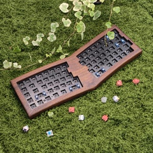 May include: A split-layout mechanical keyboard case made of dark brown wood with a black switch plate. The keyboard features several blue mechanical switches installed in the sockets, while loose pink, white, and blue switches are scattered nearby.