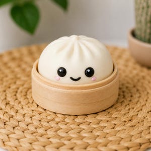 Puede incluir: Una figura de bollo al vapor blanca, redonda y de textura lisa, que presenta una cara sonriente pintada a mano en negro y pequeñas mejillas rosadas. El bollo está colocado dentro de un pequeño recipiente de bambú circular.