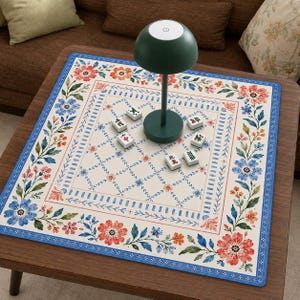 May include: A square game mat with a cream-colored center featuring a blue diamond grid pattern and scattered Mahjong tiles. The border displays a vibrant floral motif in shades of red, orange, and blue. A dark green mushroom-shaped lamp sits centrally.