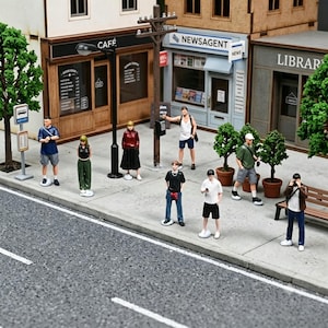 以下が含まれることがあります： カフェ、ニュースエージェント、図書館と書かれた店舗の前の歩道に立つ人々の小さなフィギュアが並ぶ、街並みを再現したミニチュアジオラマ。電柱、バス停の標識、鉢植えの木々も配置されている。