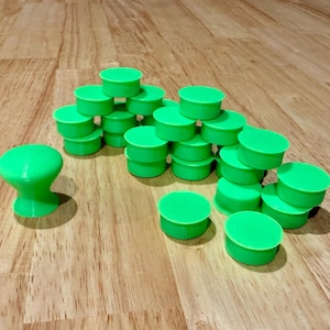 May include: A collection of bright neon green plastic components, including one standing knob with a flared base and a rounded top, alongside numerous flat, circular discs arranged in a cluster on a light-colored wooden surface.