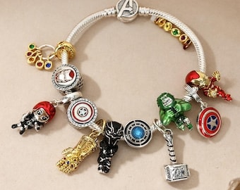 Puede incluir: Una pulsera de dijes plateada con varios colgantes de superhéroes, incluyendo un guantelete dorado, un martillo plateado, una figura verde, una máscara negra, un escudo y un cierre con el logo de los Avengers y piedras de colores.