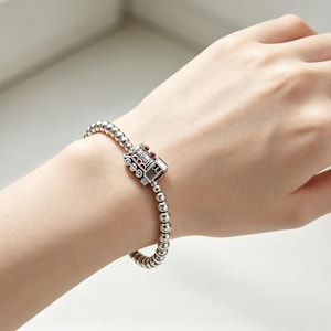 May include: A silver-toned beaded bracelet featuring a central charm shaped like a locomotive train. The train charm is detailed with small red accents on its top and side sections. The bracelet consists of a continuous string of small, polished silver-colored metal beads that form a circular band around the wrist.