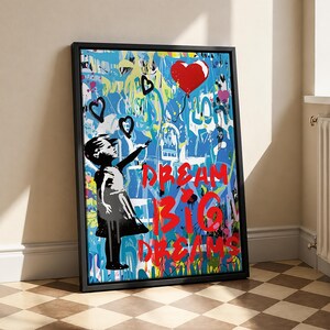 May include: A rectangular framed wall art piece featuring a black silhouette of a person reaching toward a red heart-shaped balloon. The background is a vibrant blue graffiti-style collage with the red text DREAM BIG DREAMS printed across it.