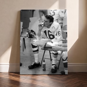 May include: A black and white photograph of a football player wearing a jersey with the number 16, sitting on a folding chair in a locker room. The player is holding a cigar to their mouth, with a glass bottle placed on the floor between their feet.