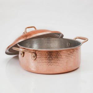 Pode incluir: Uma panela redonda de cobre martelado com duas alças laterais e uma tampa a condizer. O exterior apresenta um acabamento texturizado com covinhas num tom de cobre metálico quente, enquanto o interior é revestido com uma superfície metálica lisa prateada.