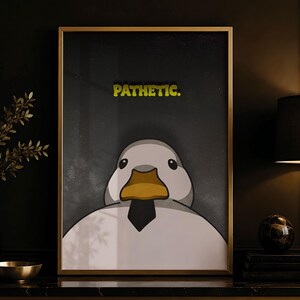 Könnte beinhalten: Ein gerahmter Kunstdruck, der eine weiße Ente mit gelbem Schnabel und schwarzer Krawatte zeigt. Der Text "PATHETIC." ist in Gelb über dem Kopf der Ente auf dunkelgrauem Hintergrund gedruckt. Der Rahmen ist dünn und goldfarben.