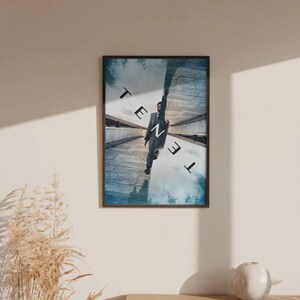May include: A framed film poster for Tenet featuring a person in a grey suit hanging from a concrete structure against a blue sky. The word TENET is printed in large black and white letters across the centre, with the image split into two mirrored perspectives showing the sky and the ground.