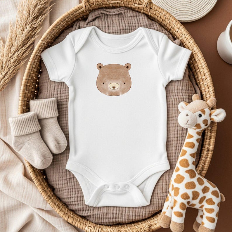 May include: White baby onesie with a brown bear face graphic. The onesie is displayed in a woven basket with a pair of beige socks and a stuffed giraffe. The background is a soft brown fabric.