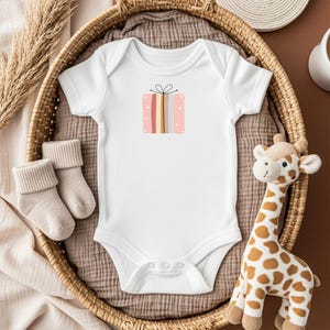 May include: White baby onesie with a colorful gift box graphic. The onesie is placed in a woven basket with a pair of beige baby socks and a stuffed giraffe toy. The gift box design features pink, yellow, and blue stripes.