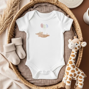 May include: White baby onesie with a watercolor illustration of a dachshund dog floating with balloons. The onesie is in a wicker basket with a pair of beige socks and a stuffed giraffe. A cute baby gift.