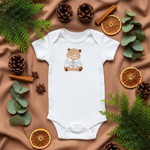 May include: White baby onesie with a cartoon bear wearing a blue jacket. The onesie is surrounded by pine cones, cinnamon sticks, and dried orange slices on a brown fabric background. A cute and cozy design for infants.