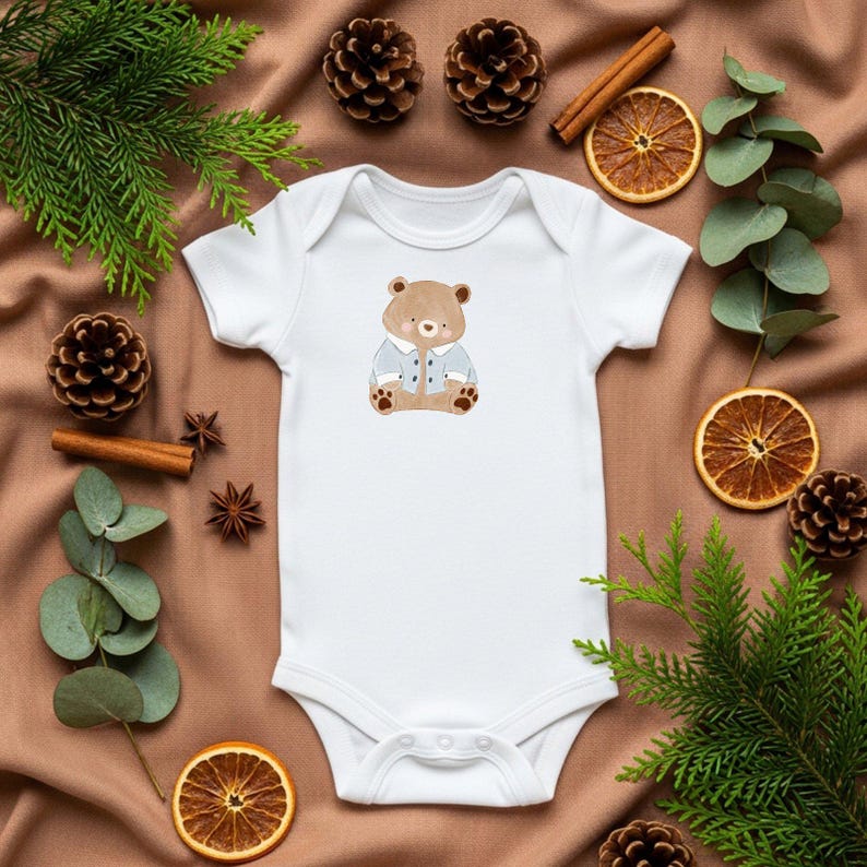 May include: White baby onesie with a cartoon bear wearing a blue jacket. The onesie is surrounded by pine cones, cinnamon sticks, and dried orange slices on a brown fabric background. A cute and cozy design for infants.