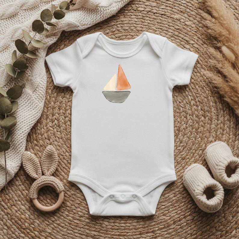 May include: White baby onesie with a watercolor sailboat design. The sailboat features an orange and cream sail atop a gray boat. A wooden bunny teether and beige baby booties are also visible on a woven mat.
