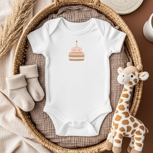 May include: White baby onesie with a small illustration of a two-tiered cake with a lit candle. The onesie is in a woven basket with a pair of beige socks and a stuffed giraffe. The background is a neutral brown.