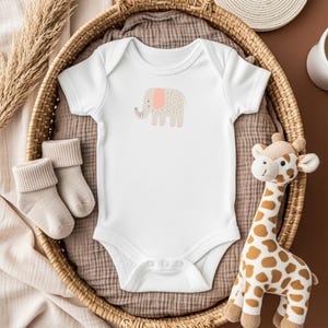 May include: A white baby onesie with a cartoon elephant illustration in beige and pink. The onesie is in a wicker basket with a pair of beige baby socks and a giraffe plush toy. The background is a neutral brown.