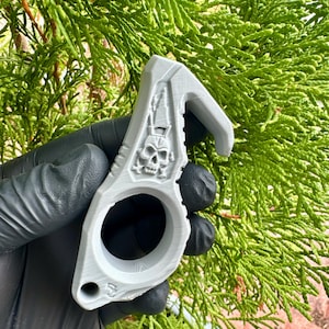Könnte beinhalten: Ein hellgraues, 3D-gedrucktes Selbstverteidigungswerkzeug mit einem Fingerloch, einer hakenförmigen Oberkante und einem erhabenen Totenkopf-Emblem auf der Vorderseite. Das Objekt hat eine matte, texturierte Oberfläche mit sichtbaren Druckschichten.