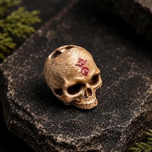 Könnte beinhalten: Eine kleine, beige, totenkopfförmige Perle mit einer verwitterten, strukturierten Oberfläche. Auf der Oberseite des Schädels befindet sich ein kleines, rotes, sternförmiges Symbol. Die Perle hat hohle Augenhöhlen, eine Nasenhöhle und ein Loch.