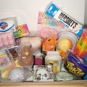Könnte beinhalten: Eine Sammlung verschiedener in Plastik verpackter Squishy-Spielzeuge, darunter ein Hershey's Cookies 'n' Creme Riegel, eine Takis-Tüte, ein Butterstück, ein Panda, ein brauner Bär, eine rosa Schokoladentafel, eine gelbe Frucht und ein Squeeze-Behälter.