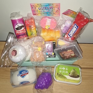 Pode incluir: Uma coleção de vários brinquedos squishy em embalagens plásticas, incluindo uma lata de Pringles, um tablete de manteiga, uma vaca highland marrom, um cacho de uvas roxas e um recipiente de sorvete de chá verde com texto em chinês e inglês.