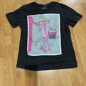 Könnte beinhalten: Ein schwarzes Kurzarm-T-Shirt mit gesprenkelter Textur und einem rechteckigen Grafikdruck des Pink Panther, der einen Pinsel und einen Farbeimer hält. Die Grafik hat einen hellgrauen Hintergrund mit einem rosa vertikalen Streifen.