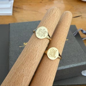 May include: Two gold signet rings with round faces, each featuring a custom engraved monogram. The rings are displayed on two light-coloured wooden ring mandrels, showcasing the polished metal finish and the intricate script lettering on each face.