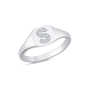 Può includere: Un anello a sigillo color argento con una parte frontale quadrata dagli angoli arrotondati. Al centro della parte frontale è presente la lettera S, impreziosita da piccole pietre chiare e scintillanti incastonate nella superficie metallica lucida.