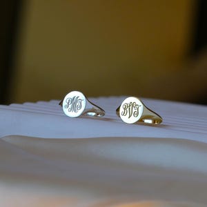 Pode incluir: Dois anéis de sinete com faces circulares, um em tom prateado e outro em tom dourado. Cada anel apresenta um monograma gravado na superfície circular plana. O anel prateado exibe as letras PMT, enquanto o anel dourado exibe as letras BVJ.