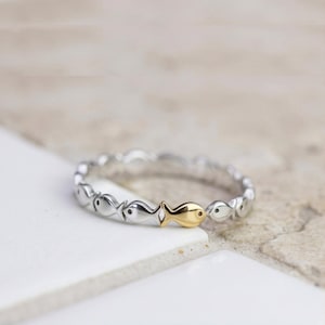 May include: A silver-toned ring featuring a continuous band of small, stylized fish shapes linked together. One single fish in the center of the band is finished in a contrasting gold tone, while all other fish are silver with small black dots for eyes.