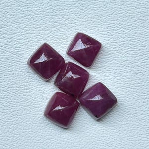 Könnte beinhalten: Fünf quadratisch geschliffene, tief magentafarbene Edelsteine, die in einer Gruppe angeordnet sind. Jeder Stein weist eine facettierte, pyramidenartige Oberfläche mit einem glatten, polierten Finish auf, das das Licht reflektiert.