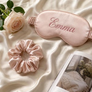 May include: A pale pink satin sleep mask embroidered with the name Emma in a matching thread, accompanied by a matching pale pink satin scrunchie. The items are displayed alongside a pale pink rose and an open book with a floral illustration.