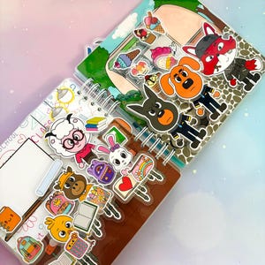 Peut inclure: Un carnet à spirales orné d'une collection d'autocollants de personnages de dessins animés colorés. Les autocollants représentent des animaux comme un mouton, un lapin, un chien et un renard, ainsi que des objets scolaires, livres et glaces.