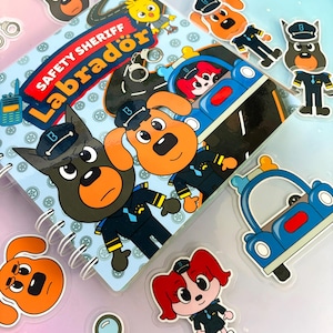 Puede incluir: Un cuaderno de espiral titulado Safety Sheriff Labrador con perros policías de dibujos animados en uniformes azules, un coche de policía azul y una lupa. La portada incluye ilustraciones de perros con gorras, esposas y un personaje de pollito amarillo.