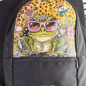 Pode incluir: Uma mochila preta com um grande gráfico de um sapo verde usando óculos de sol cor-de-rosa e um colar de contas. O fundo do gráfico tem um padrão de leopardo, flores, penas e o texto DARE TO DREAM. O logótipo da adidas é branco.
