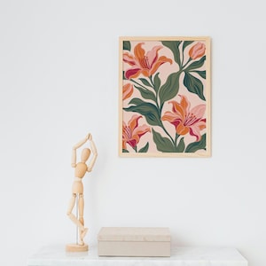 Peut inclure: Une impression murale verticale encadrée présentant un motif répétitif de lys orange et roses avec un feuillage vert foncé sur fond rose clair, accompagnée d'un mannequin d'artiste en bois et d'une boîte rectangulaire beige.