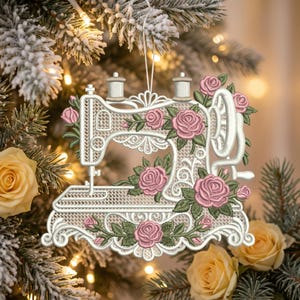 May include: A white embroidered sewing machine ornament featuring intricate lace-like patterns and detailed pink roses with green leaves. The design includes two small thread spools at the top and is suspended by a thin white string for hanging.