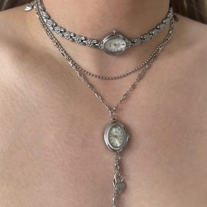 Könnte beinhalten: Ein mehrlagiges silberfarbenes Halskettenset mit zwei Uhrenzifferblättern. Die obere Lage ist ein Choker mit einer kleinen ovalen Uhr, während die untere Lage eine längere Kette mit einer größeren ovalen Uhr und zwei kleinen herzförmigen Anhängern ist.