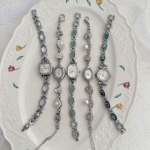 Op de afbeelding: Vijf zilverkleurige polshorloges met decoratieve schakelarmbanden. De horloges zijn voorzien van diverse accenten, waaronder ovale lichtblauwe stenen, witte parels, groene ovale cabochons, heldere strass-steentjes en inlays van abalone-schelp.