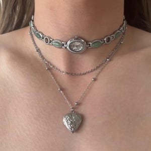 May include: A layered necklace set featuring a silver-toned watch choker with oval light green stone accents and a delicate chain necklace with a silver heart-shaped locket pendant featuring an embossed floral design on the front surface.