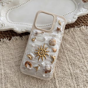 May include: A beige smartphone case featuring a textured white surface decorated with various gold-toned metal charms including a sun, a dolphin, a crab, and a mermaid tail, alongside several small natural seashells and white pearl-like beads.