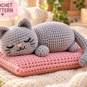 Puede incluir: Un gato de ganchillo gris con los ojos cerrados, una pequeña nariz rosa y bigotes bordados, descansando sobre un cojín rectangular de ganchillo rosa. Una etiqueta rosa en la esquina superior izquierda muestra el texto CROCHET PATTERN.